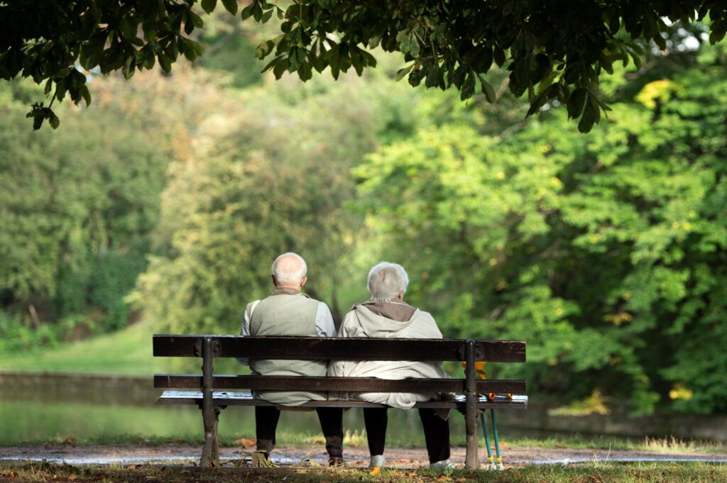 Zwei Senioren sitzen mit dem Rücken zur Kamera auf einer Parkbank in einem grünen Parkanlage.
