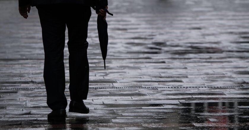 Ein Passant geht mit einem Regenschirm in der Hand über einen nassen Gehweg.