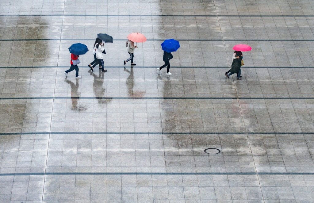 Wenige Menschen laufen bei Regenwetter mit Regenschirmen über einen offenen Platz.