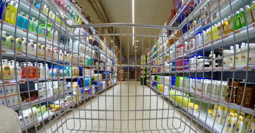 Ein Supermarkt aus der Sicht eines Einkaufswagens. Links und Rechts sind Produkte in vollen Regalen.