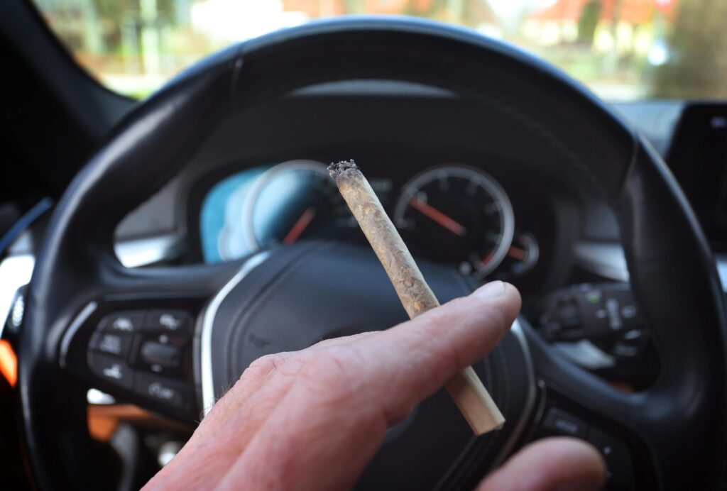 Eine Hand hält einen Joint im Auto vor einem Lenkrad.