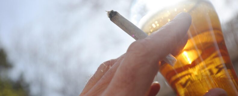 Eine Zigarette in der Hand und ein Bier in Nahaufnahme vor einem blauen Himmel.